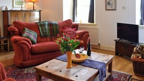 A living room with a red sofa, a wooden coffee table with a vase of flowers, a television, and a fireplace. - 2 bedroom accommodation in Town Yetholm, near Kelso (Town Yetholm, near Kelso)