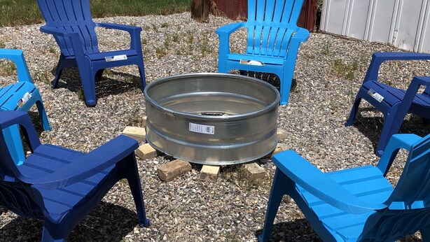 A circular metal fire pit surrounded by six blue plastic Adirondack chairs arranged in a circle on a gravel surface.