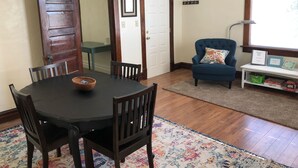 A dining area with a round table, four chairs, a wooden door, a blue armchair, and a small white console table with a basket and picture frames.