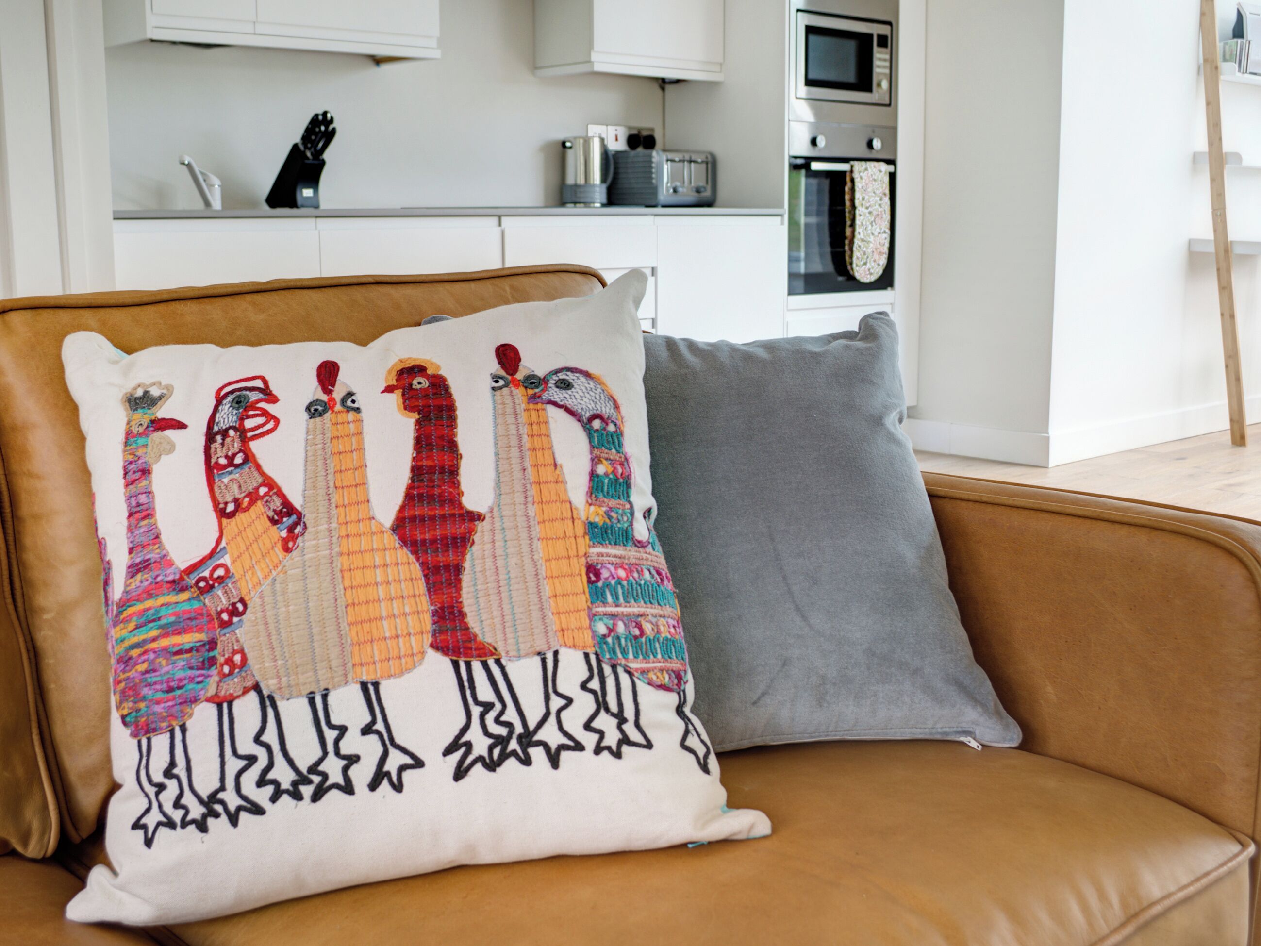 A brown leather sofa with a colorful embroidered pillow depicting birds and a grey pillow.