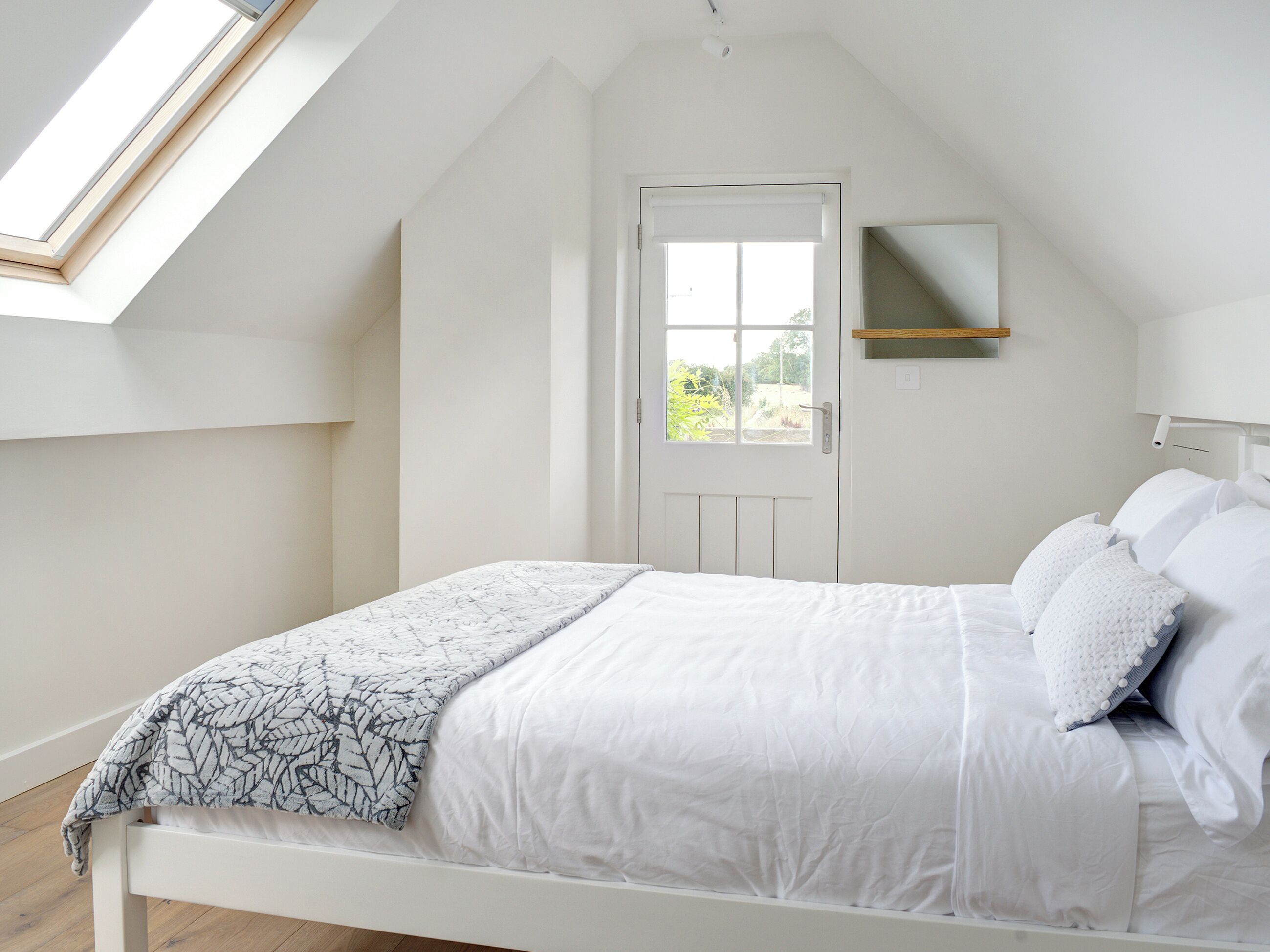 A bedroom with a sloped ceiling, a skylight, a white door, and a bed with white bedding and pillows.