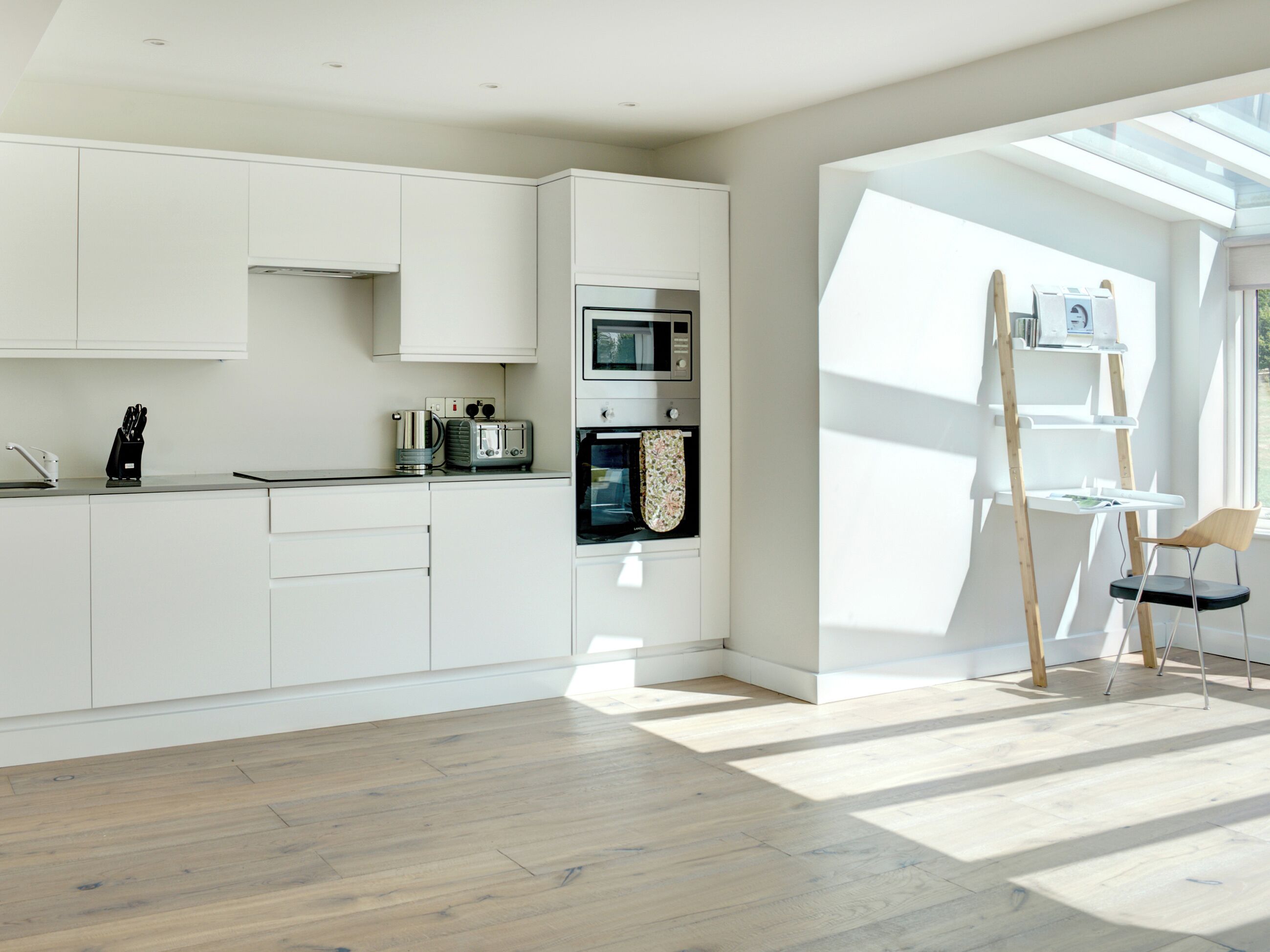 Een moderne keuken met witte kasten, een magnetron, oven en een broodrooster. Er staat een houten ladder en een stoel bij een raam.
