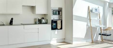 Een moderne keuken met witte kasten, een magnetron, oven en een broodrooster. Er staat een houten ladder en een stoel bij een raam.