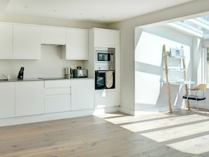 Une cuisine moderne avec des meubles blancs, un four à micro-ondes, un four et un grille-pain. On y trouve une échelle en bois et une chaise près d’une fenêtre.