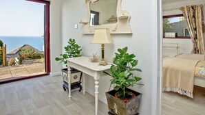 Ein Zimmer mit großem Fenster, einem Bett, einem Nachttisch mit Lampe, einem dekorativen Spiegel und einer Topfpflanze.