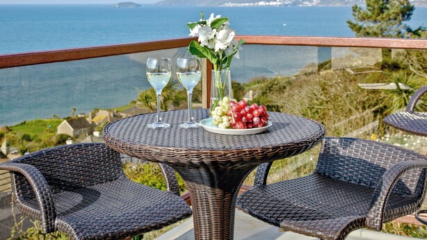 Ein Korbstuhl mit zwei Gläsern Weißwein, einer Blumenvase und einer Platte mit Trauben auf einem Balkon mit Blick auf das Meer.