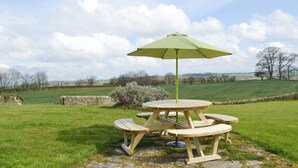 A picnic table with a green umbrella in a grassy field with stone walls and trees in the background.