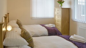 A bedroom with two beds, a wooden dresser, a window with blinds, and a vase with flowers on top of the dresser.