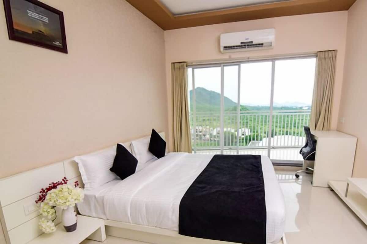 A hotel room with a bed, a bedside table with a vase of flowers, a chair, and a large window with a view of a mountain and buildings.