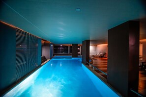 A modern indoor swimming pool with a glass wall on one side, a wooden deck area, and a seating area with chairs. - G G Holiday Homes Innovating Hospitality (Udaipur)