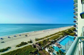 A beach view from a hotel balcony with a swimming pool and beach umbrellas.