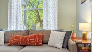 A living room with a beige sofa, patterned cushions, a window with patterned curtains, a small table with a lamp, and a bookshelf with board games.