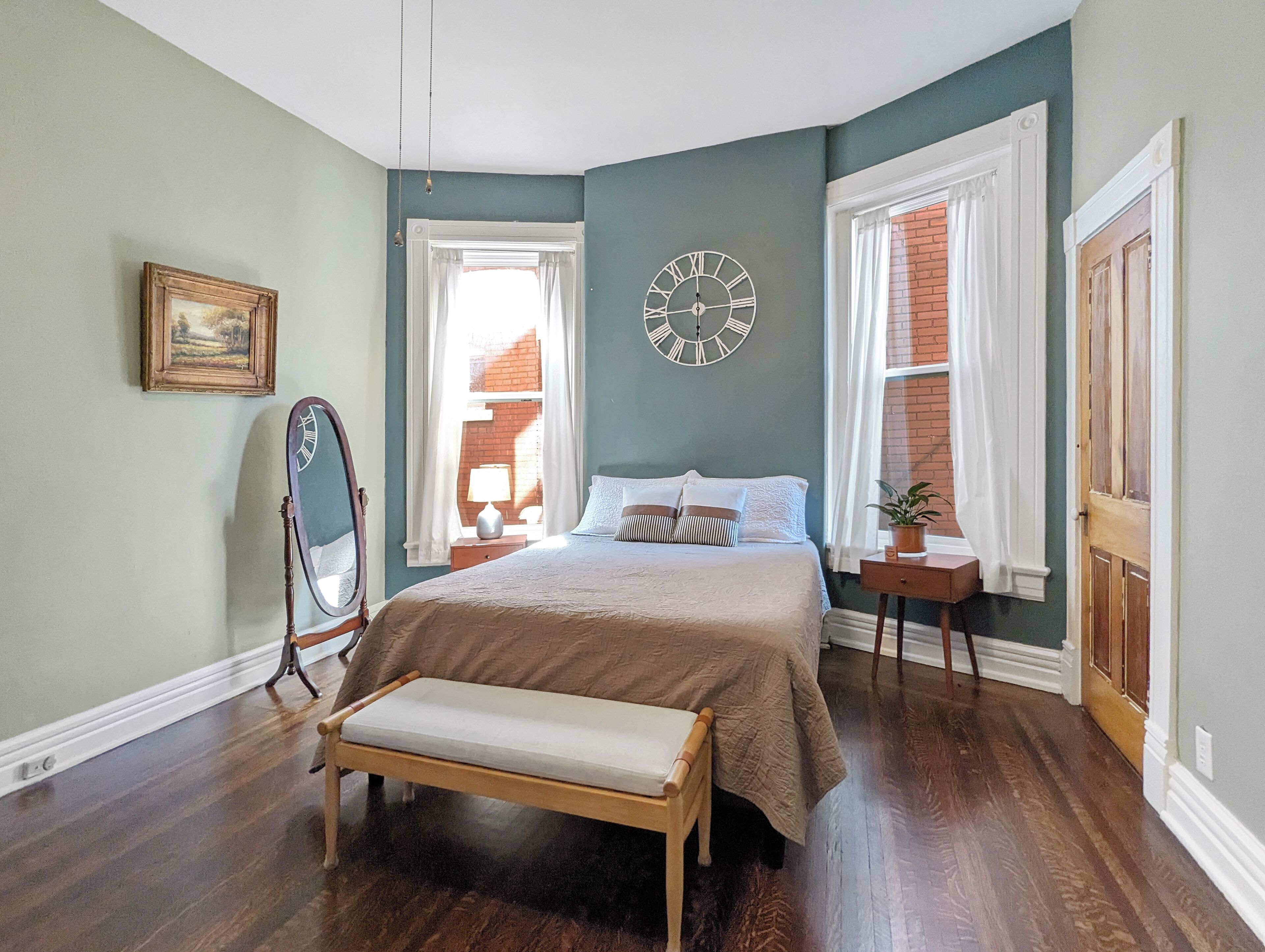 Un dormitorio con cama, un banco, un espejo, un reloj y una ventana con cortinas.