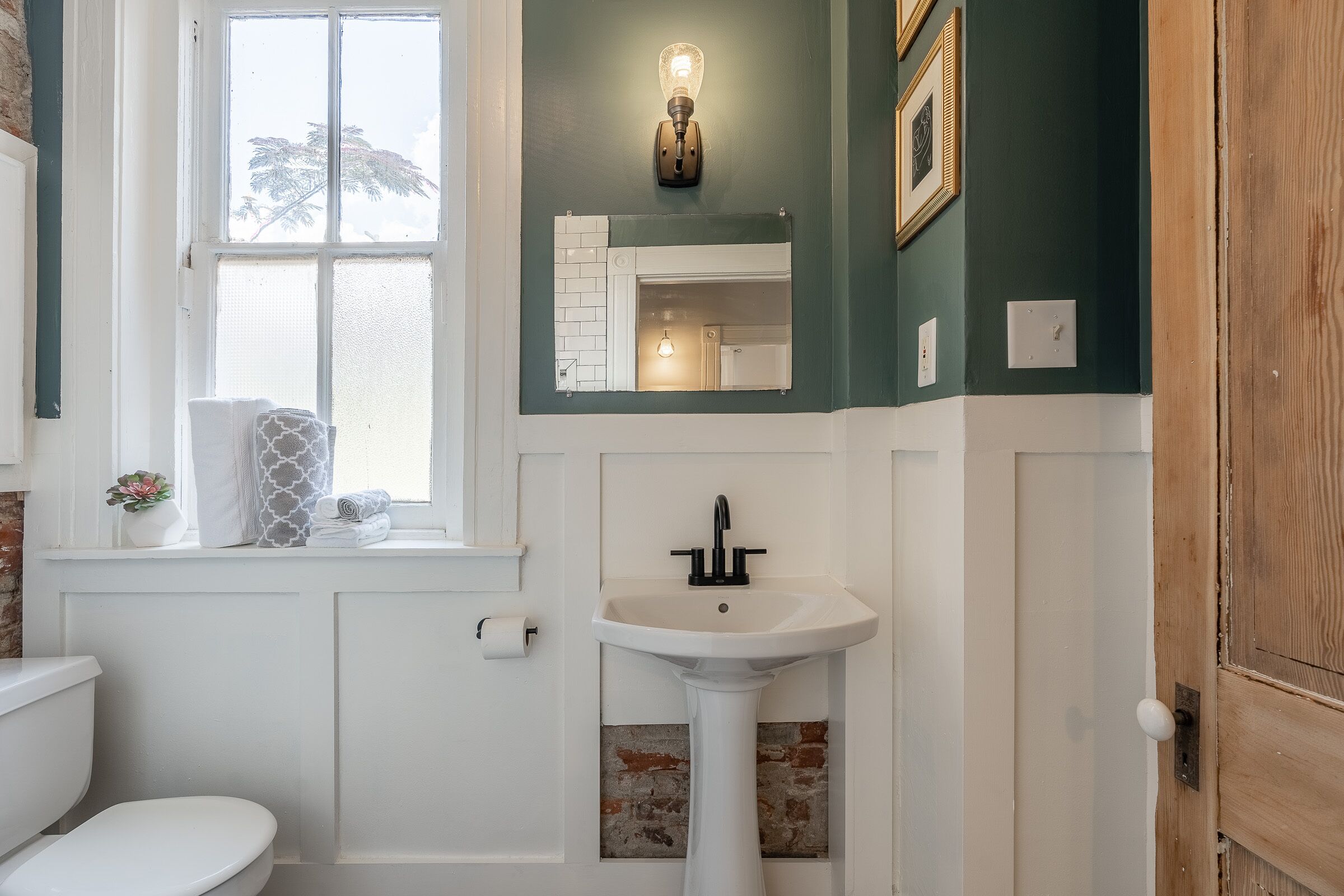 A bathroom with a white toilet, a pedestal sink, a window with a view of trees, and a mirror above the sink.