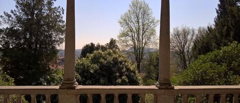 Une balustrade en pierre, ornée de colonnes, surplombe un paysage composé d’arbres et de collines lointaines.