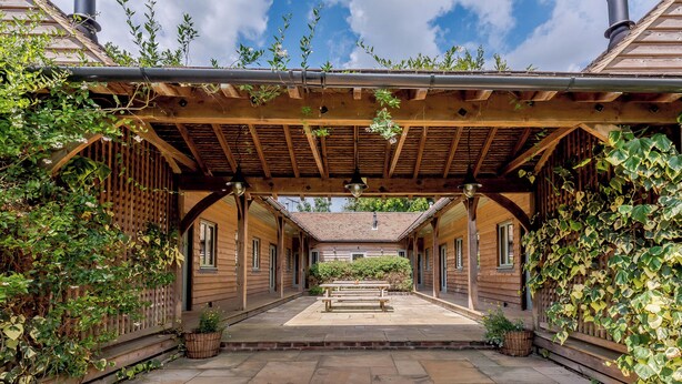 A covered outdoor patio with wooden beams and a stone floor, surrounded by wooden buildings and greenery. - Castlemans Stables West (Sedlescombe, near Battle)