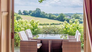 An outdoor patio with wicker furniture and a glass table, overlooking a green landscape with trees and a distant house. - Castlemans Stables West (Sedlescombe, near Battle)