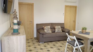 Uma sala de estar aconchegante com um sofá marrom, duas almofadas decorativas, uma mesa lateral de madeira com uma planta e uma cadeira branca.