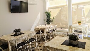 Uma sala de jantar com mesas e cadeiras, uma televisão de tela plana presa na parede e uma janela grande que ilumina naturalmente o espaço.
