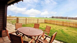 Une terrasse extérieure avec une table et des chaises en bois, une clôture en bois et, au fond, un champ d’herbe.