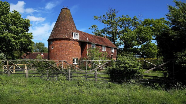 A brick tower house with a conical roof, surrounded by lush greenery and a wooden fence.