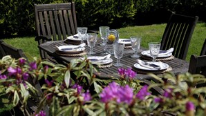 An outdoor dining table set with plates, glasses, and cutlery, surrounded by chairs and a hedge.