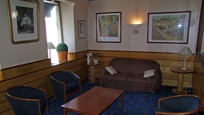 Een kamer met een bruine bank, twee blauwe fauteuils, een houten salontafel en een klein bijzettafeltje met een lamp. Er hangen ingelijste foto’s aan de muur en een bloempot op de vensterbank.