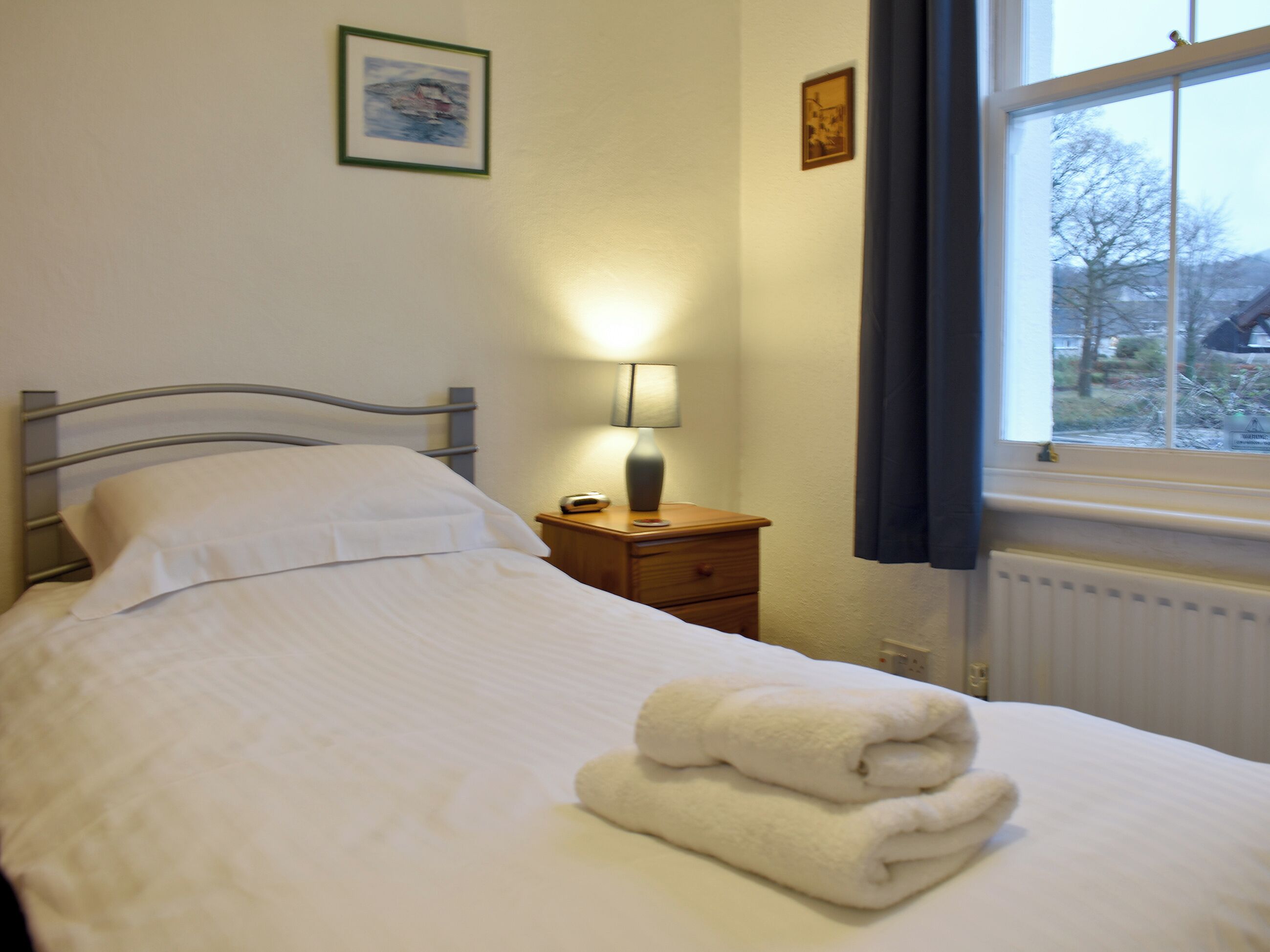 Un lit bien fait, avec du linge de lit blanc et une serviette pliée posée sur le côt é. Il y a une table de chevet avec une lampe, et un tableau encadré est accroché au mur juste au-dessus.