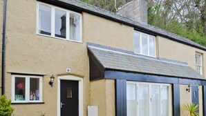 Une maison à deux étages, à la façade beige, avec une porte noire et un petit jardin devant, composé de gravier et d’une plante en pot.