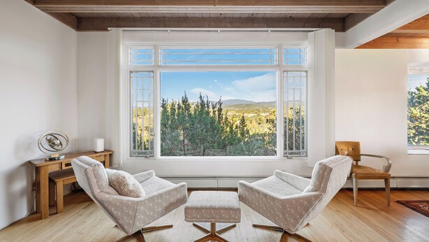 A modern living room with two armchairs, a small ottoman, and a wooden coffee table. A large window offers a view of a forest and mountains. - Heavenly Mountain Views-Close to Plaza, Hot tub, Ski & Pickleball, Upscale home (Santa Fe)