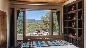 A bedroom with a large window, a bed, a wooden headboard, a wooden cabinet with decorative items, and a view of a landscape with trees and mountains. - Heavenly Mountain Views-Close to Plaza, Hot tub, Ski & Pickleball, Upscale home (Santa Fe)