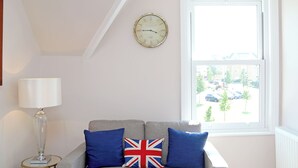 Ein Zimmer mit einem grauen Sofa, zwei blauen Kissen mit Union-Jack-Motiv, einer runden Wanduhr und einem Fenster mit Blick auf geparkte Autos und Gebäude.