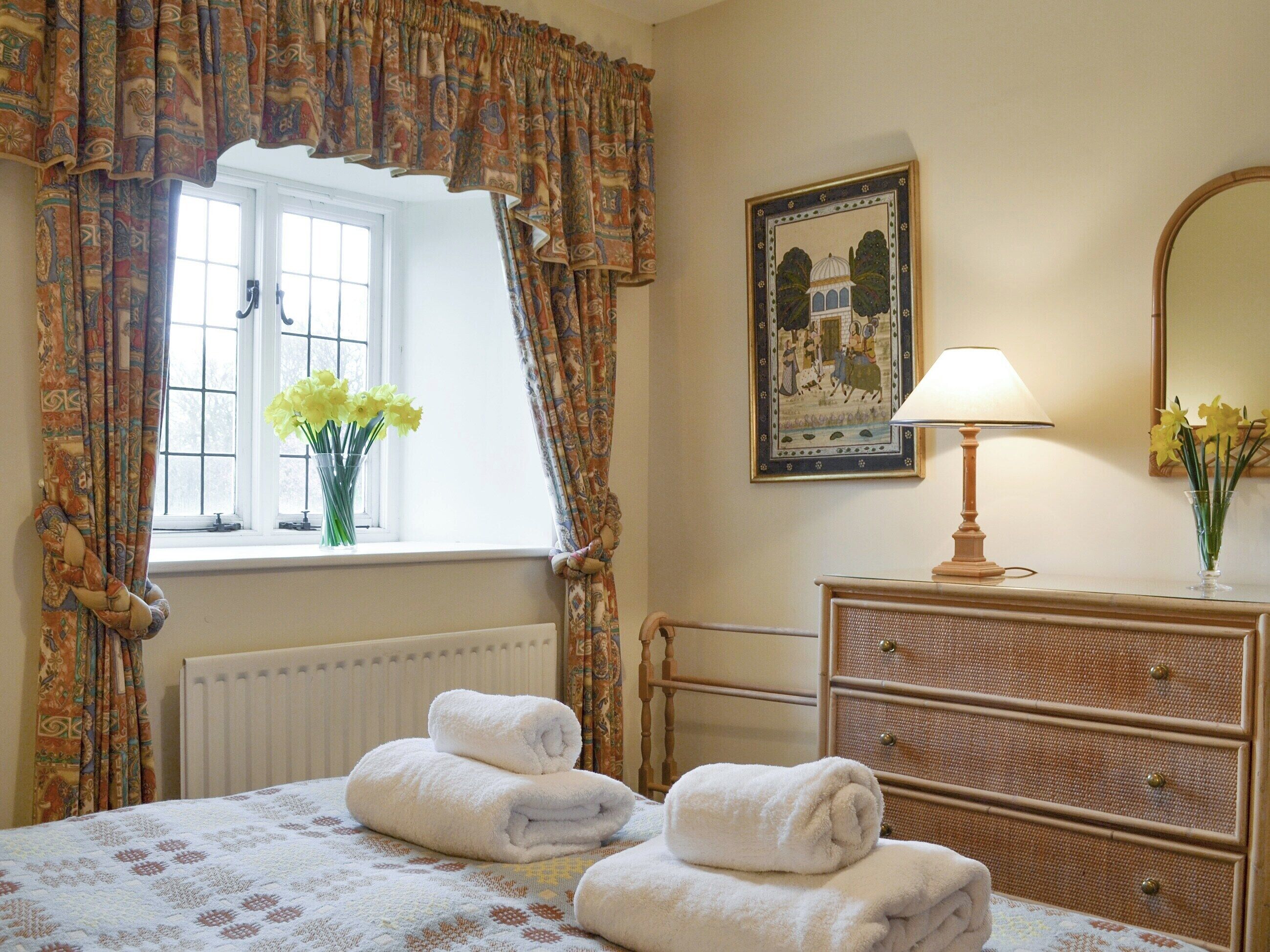 A hotel room with a bed, a dresser, a window with curtains, a radiator, a lamp, and a framed picture on the wall.