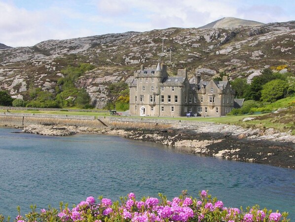 A castle-like building with multiple towers, situated by a body of water with rocky terrain in the background.