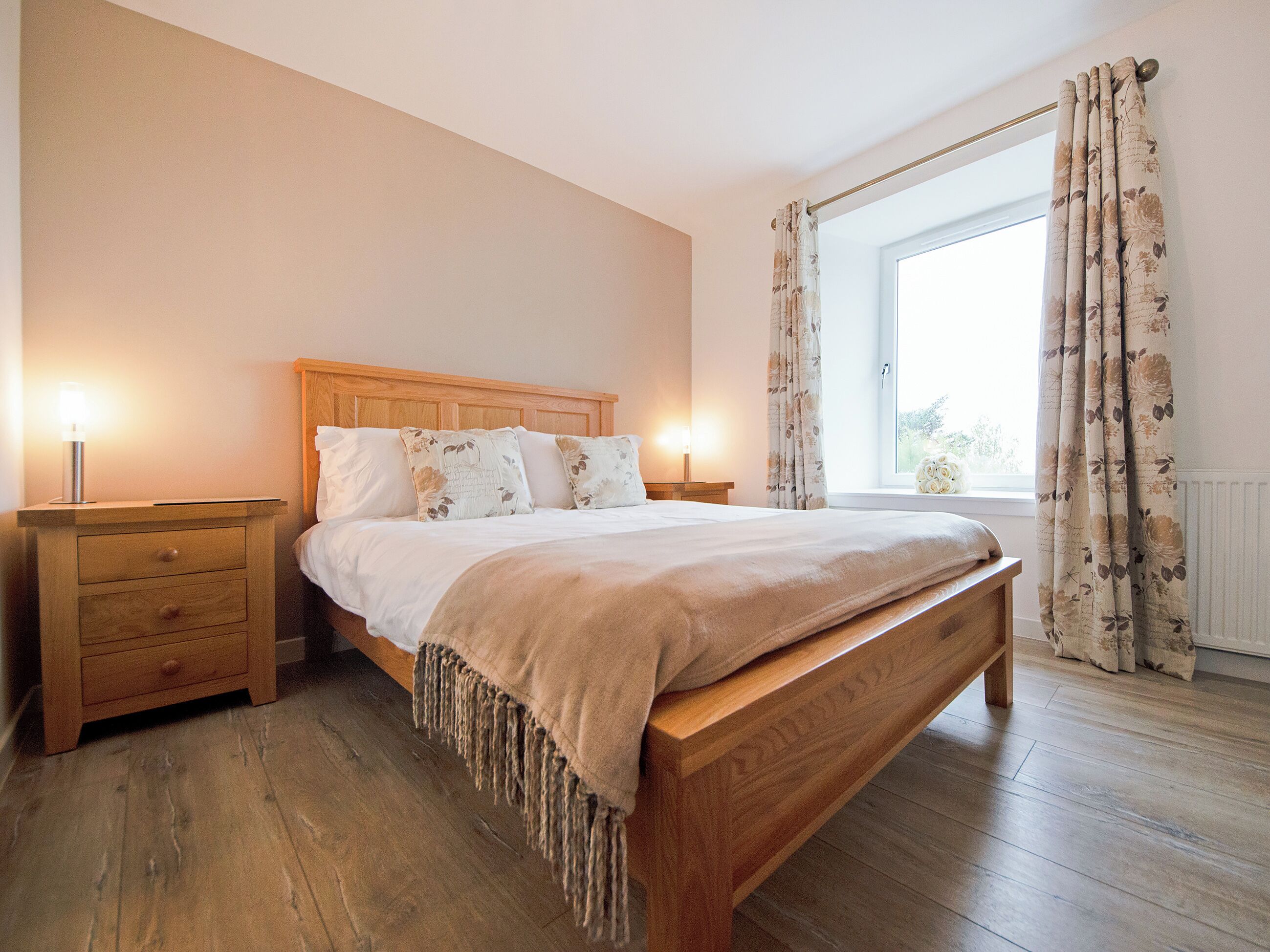 Quarto com cama de madeira, duas mesas de cabeceira, janela com cortinas e radiador.