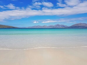 A beach with clear turquoise water, a sandy shore, and distant mountains under a blue sky.