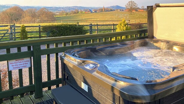Una vasca idromassaggio su un pontile in legno con vista su un campo e edifici in lontananza.