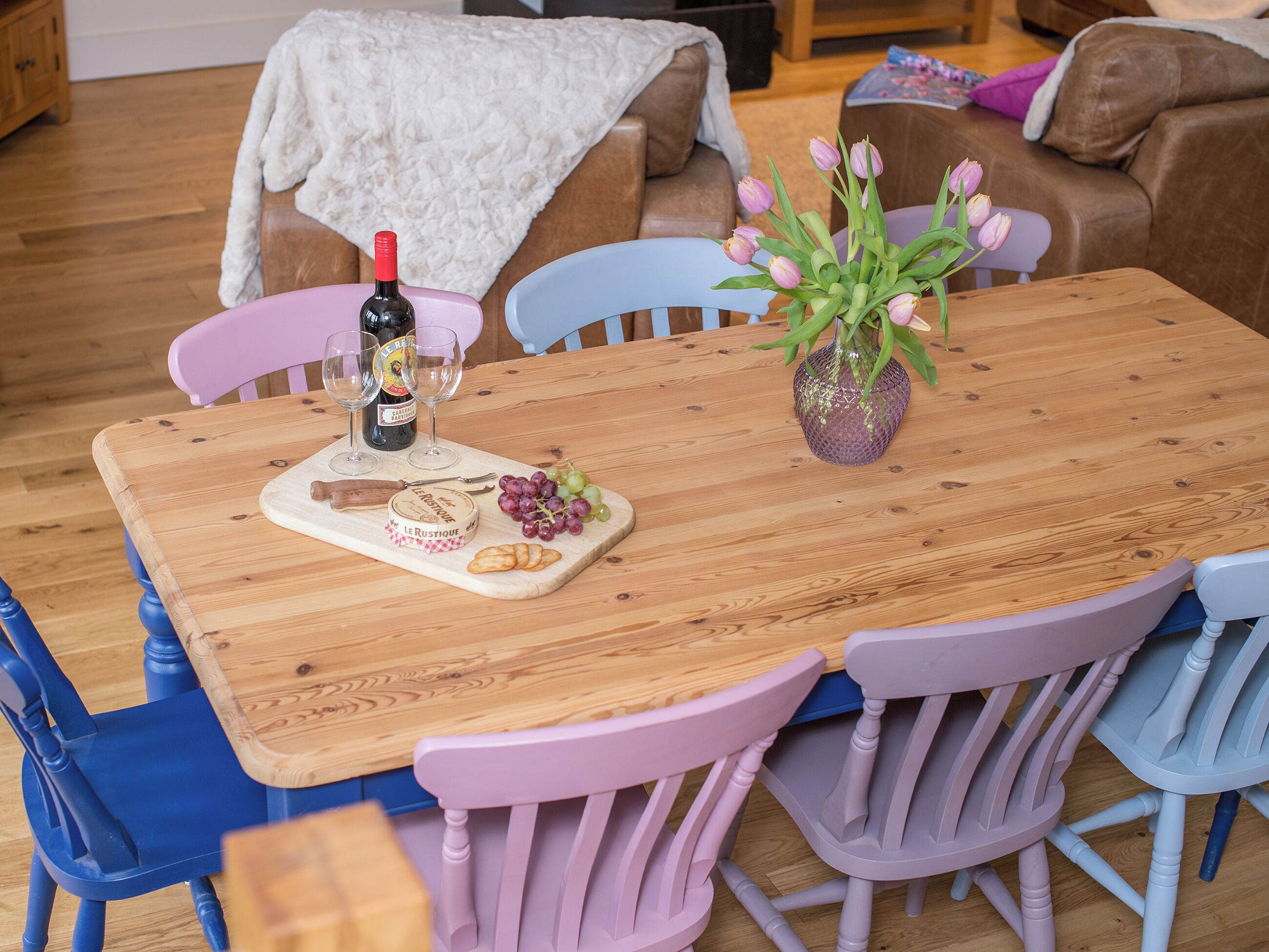 A wooden dining table with a bottle of wine, glasses, cheese, grapes, and a vase of flowers.