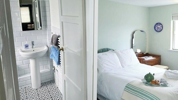 Camera d'albergo con bagno dotato di lavandino e specchio bianchi, pavimento decorato e un letto con biancheria da letto bianca, più un comodino con lampada e un piatto di cibo.