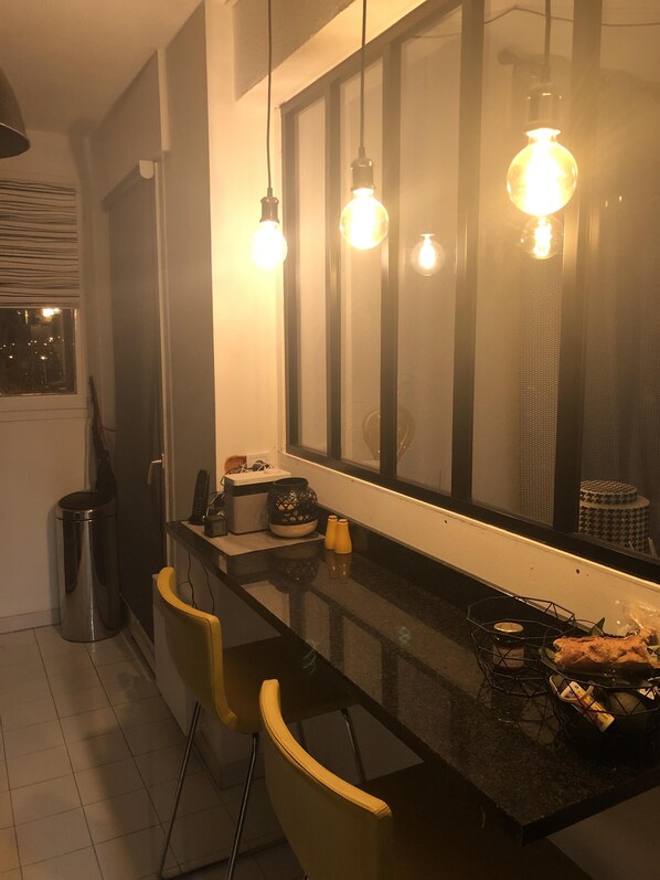 Una cocina con un espejo grande, lámparas colgantes, un cubo de basura, un refrigerador, una mesa pequeña con un tazón y una silla.