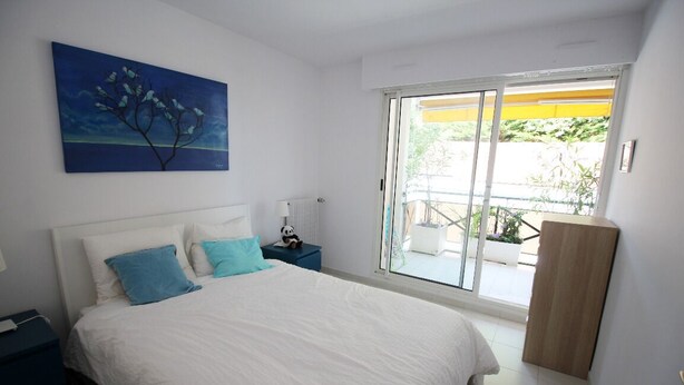 Un dormitorio con una cama grande, una mesita de noche, una lámpara, un cuadro y un ventanal corredera que da a un espacio exterior.