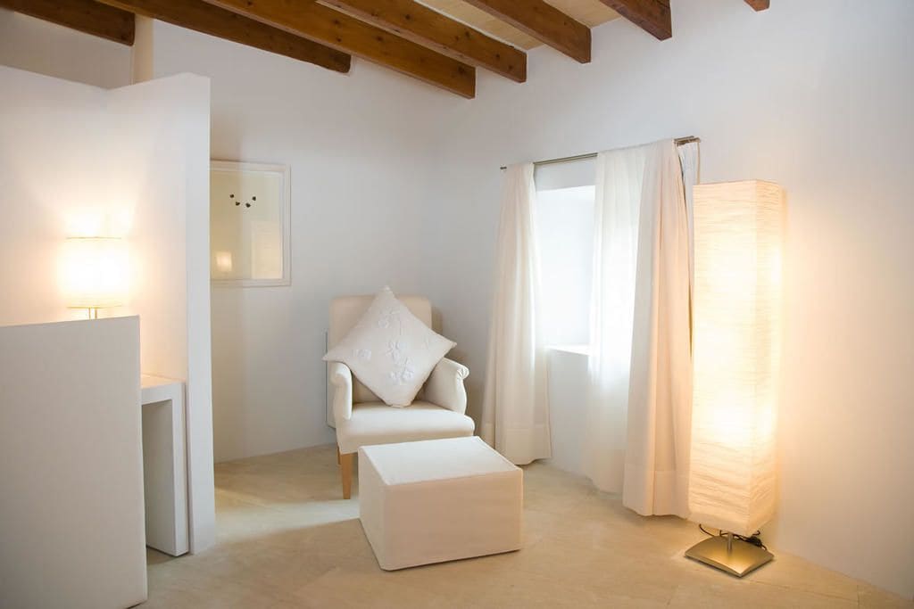 Una habitación minimalista con un sillón blanco, un pequeño reposapiés, una lámpara de pie y una ventana con cortinas.