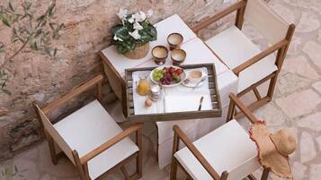 Una pequeña mesa al aire libre con una bandeja de comida y bebidas, rodeada de sillas de madera con cojines blancos.