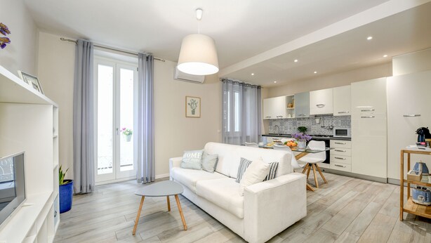 A modern living room with a white sofa, a small round table, and a TV. There is a kitchen area with cabinets and a dining table. - 50 meters from the lake, in the historic center with a terrace. Stella House (Desenzano del Garda)