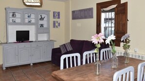 A living room with a TV, grey cabinets, a purple sofa, a dining table with chairs, and a window with a view. - VISTA VELETA CASA ROMERO (Capileira)