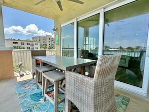 Un balcón con una mesa y sillas de mimbre, ventanas amplias y vistas a edificios.