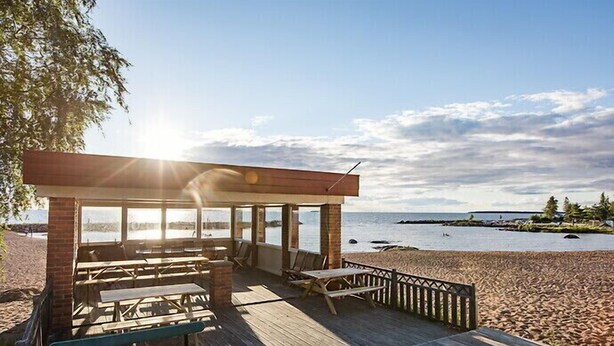 Un muelle de madera frente a la playa con mesas y sillas de picnic, con vista a un mar tranquilo.