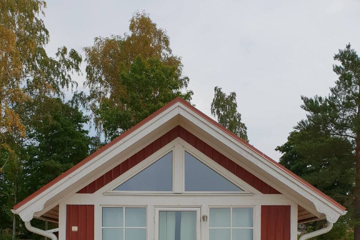 A small wooden cabin with a red exterior, a gabled roof, and a deck with two wooden chairs and a table.