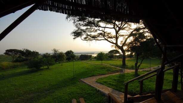 A view from a covered outdoor area with a wooden deck and chairs, overlooking a grassy landscape with trees and a distant horizon. - Bwana Tembo Safari Lodge (Pakwach)