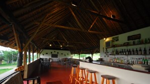 A covered outdoor seating area with wooden stools and tables, a bar with shelves of bottles, and a view of greenery outside. - Bwana Tembo Safari Lodge (Pakwach)